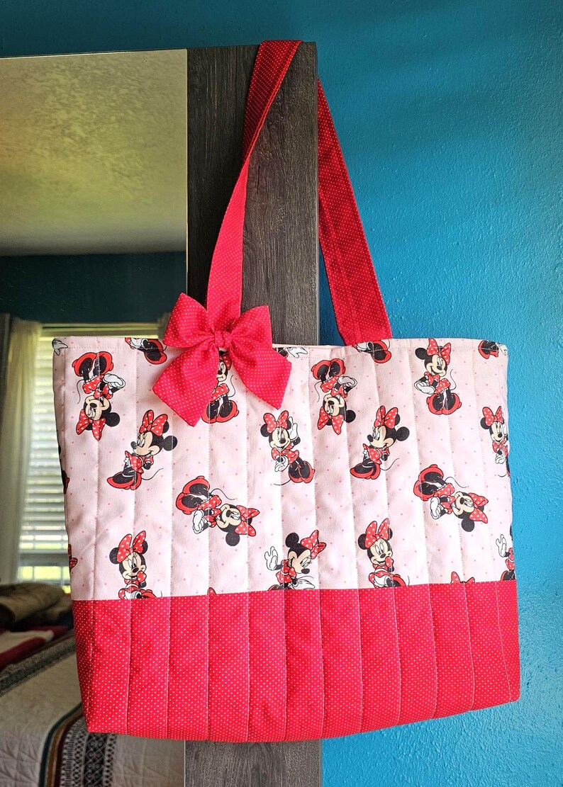 Puede incluir: Un bolso tote acolchado con un estampado de Minnie Mouse sobre un fondo rosa claro. Tiene detalles de lunares rojos, un lazo rojo y correas rojas. El bolso mide aproximadamente 38 cm de ancho y 30 cm de alto.