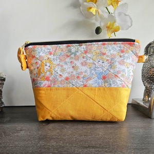 May include: A trapezoidal zippered pouch featuring a top panel with a floral print and cartoon princess characters, and a solid mustard yellow bottom panel. The pouch has quilted stitching, a black zipper, and gold-toned metal side loops.