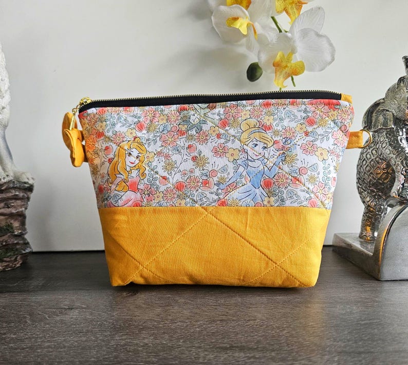 May include: A trapezoidal zippered pouch featuring a top panel with a floral print and cartoon princess characters, and a solid mustard yellow bottom panel. The pouch has quilted stitching, a black zipper, and gold-toned metal side loops.