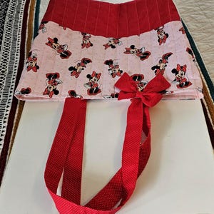 Puede incluir: Un bolso tote de Minnie Mouse rojo y rosa. El bolso presenta un lazo y correas de lunares rojos. El cuerpo principal del bolso es rosa con un estampado de Minnie Mouse. La parte superior del bolso es roja con un estampado de lunares.