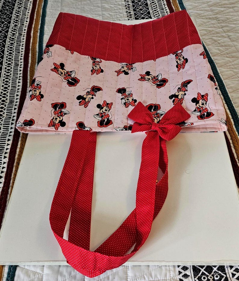 Puede incluir: Un bolso tote de Minnie Mouse rojo y rosa. El bolso presenta un lazo y correas de lunares rojos. El cuerpo principal del bolso es rosa con un estampado de Minnie Mouse. La parte superior del bolso es roja con un estampado de lunares.