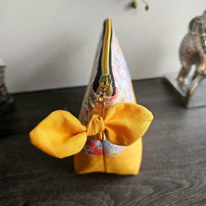 May include: A zippered pouch featuring a floral pattern in shades of blue, orange, and white on the upper section, with a solid mustard yellow base. The gold-toned zipper pull is accented with a decorative mustard yellow fabric bow.