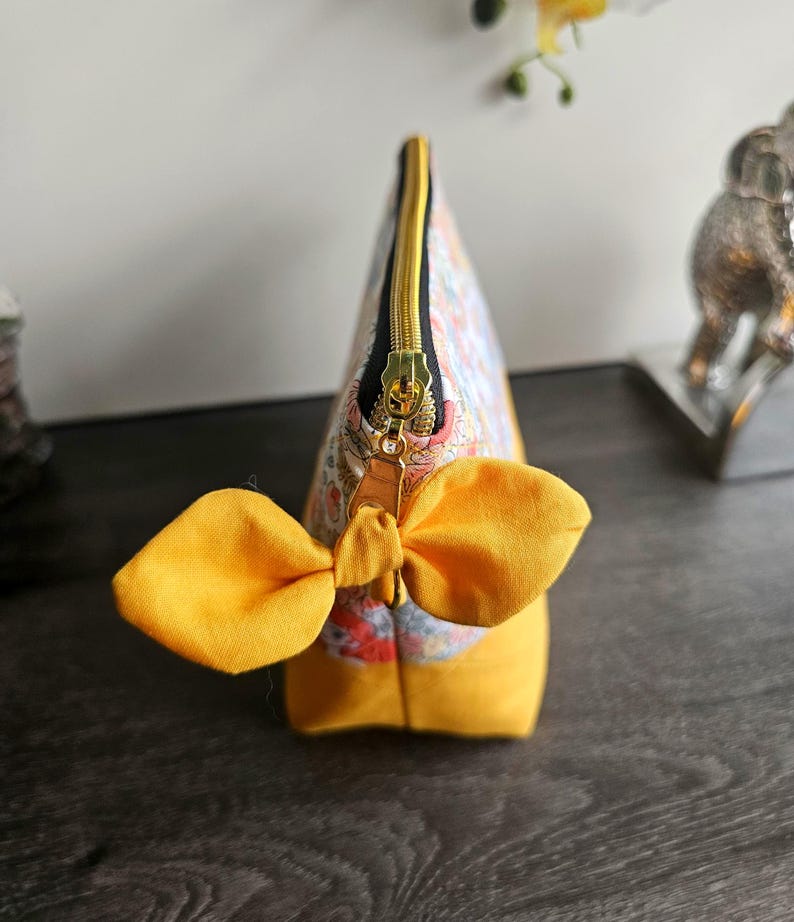 May include: A zippered pouch featuring a floral pattern in shades of blue, orange, and white on the upper section, with a solid mustard yellow base. The gold-toned zipper pull is accented with a decorative mustard yellow fabric bow.