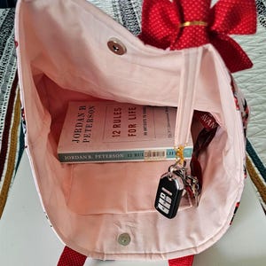 Puede incluir: Un bolso tote rosa con un lazo y correas rojas. Dentro del bolso hay un libro titulado "12 Reglas para la Vida" de Jordan B. Peterson, llaves y un cord&oacute;n. El bolso tiene un cierre magn&eacute;tico.