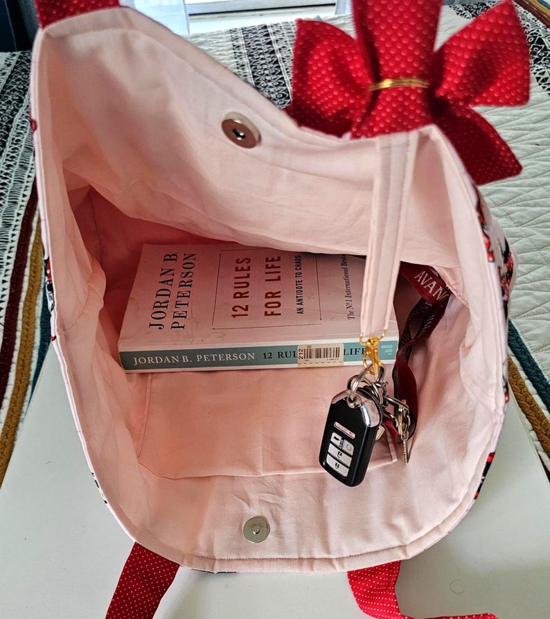 Puede incluir: Un bolso tote rosa con un lazo y correas rojas. Dentro del bolso hay un libro titulado "12 Reglas para la Vida" de Jordan B. Peterson, llaves y un cord&oacute;n. El bolso tiene un cierre magn&eacute;tico.