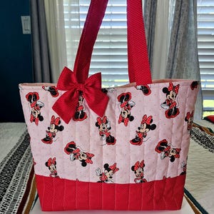 Puede incluir: Un bolso tote rosa con un dise&ntilde;o de Minnie Mouse, lazos y detalles rojos. El bolso tiene asas rojas y un lazo grande en la parte delantera. La base del bolso es roja con un estampado de lunares.