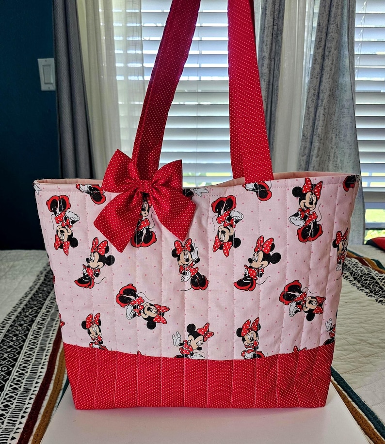 Puede incluir: Un bolso tote rosa con un dise&ntilde;o de Minnie Mouse, lazos y detalles rojos. El bolso tiene asas rojas y un lazo grande en la parte delantera. La base del bolso es roja con un estampado de lunares.