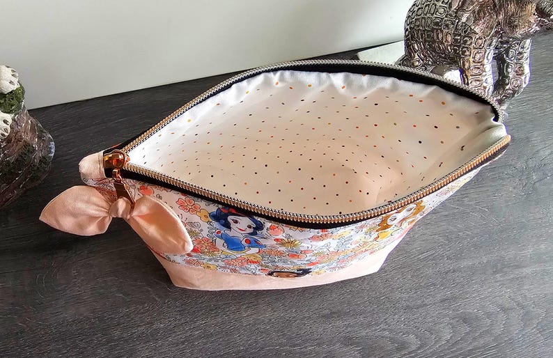 May include: A zippered pouch featuring a colorful Snow White and forest animal print on the exterior, a solid light pink base, and a light pink interior lining with small gold polka dots. The pouch includes a small pink fabric bow on the side.