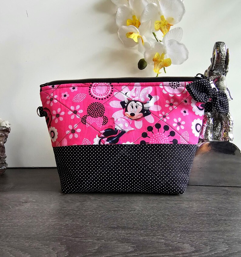 Peut inclure: Une pochette zipp&eacute;e matelass&eacute;e pr&eacute;sentant un tissu rose vif avec un motif Minnie Mouse et floral sur la moiti&eacute; sup&eacute;rieure et un tissu noir &agrave; pois blancs sur la moiti&eacute; inf&eacute;rieure. Un petit n&oelig;ud &agrave; pois noir et blanc est attach&eacute; &agrave; la fermeture.
