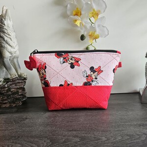 Pu&ograve; includere: Una pochette trapuntata con cerniera caratterizzata da un tessuto rosa chiaro con motivo Minnie Mouse nella parte superiore e un tessuto rosso a pois bianchi nella parte inferiore, rifinita con un piccolo fiocco in tessuto rosso sulla zip.