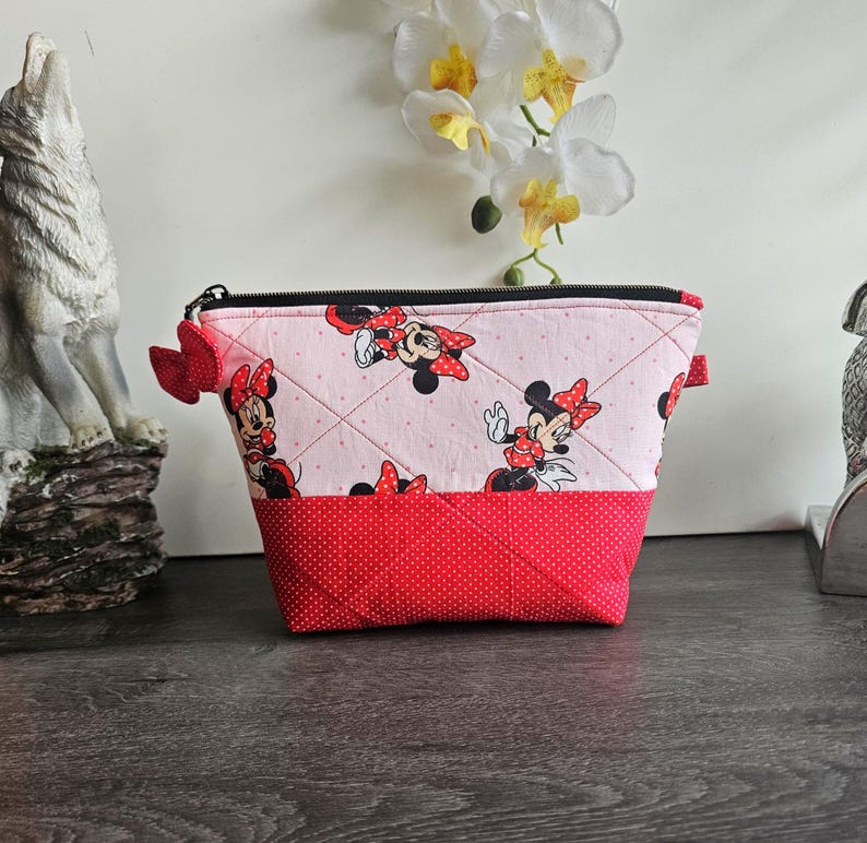 Pu&ograve; includere: Una pochette trapuntata con cerniera caratterizzata da un tessuto rosa chiaro con motivo Minnie Mouse nella parte superiore e un tessuto rosso a pois bianchi nella parte inferiore, rifinita con un piccolo fiocco in tessuto rosso sulla zip.