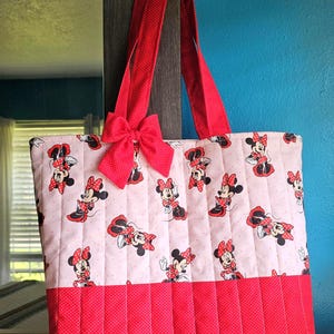 Puede incluir: Un bolso tote acolchado con un estampado de Minnie Mouse sobre un fondo rosa claro. El bolso tiene asas rojas y un lazo rojo. La parte inferior del bolso es roja con un estampado de lunares.