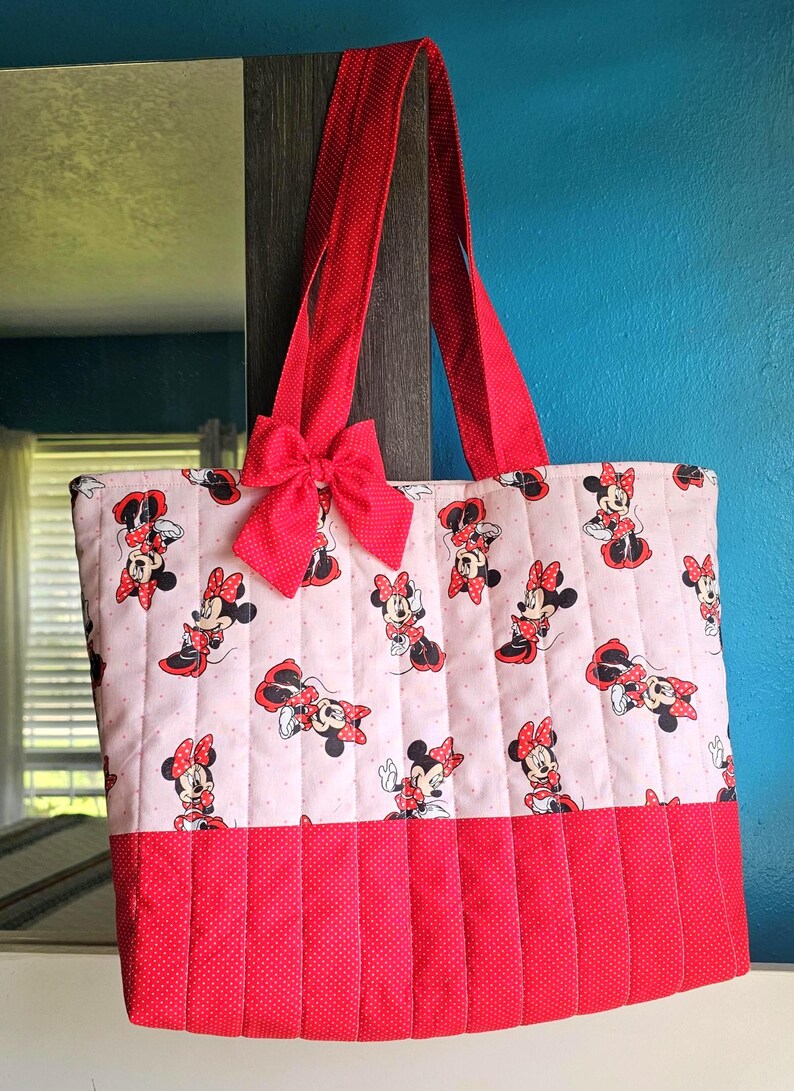 Puede incluir: Un bolso tote acolchado con un estampado de Minnie Mouse sobre un fondo rosa claro. El bolso tiene asas rojas y un lazo rojo. La parte inferior del bolso es roja con un estampado de lunares.