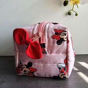 Puede incluir: Un neceser acolchado rosa con un estampado repetido de Minnie Mouse. La bolsa tiene un lazo rojo de lunares y una cremallera rosa. La bolsa tiene forma de cubo y parece estar hecha de tela.