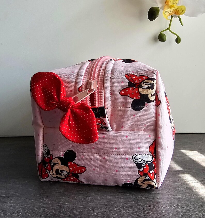 Puede incluir: Un neceser acolchado rosa con un estampado repetido de Minnie Mouse. La bolsa tiene un lazo rojo de lunares y una cremallera rosa. La bolsa tiene forma de cubo y parece estar hecha de tela.