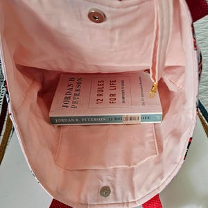 Puede incluir: Bolso tote abierto con interior rosa claro y un libro titulado "12 Reglas para la Vida" en su interior. El bolso tiene una correa de lunares rojos y un cierre magn&eacute;tico. El exterior presenta un dise&ntilde;o estampado.
