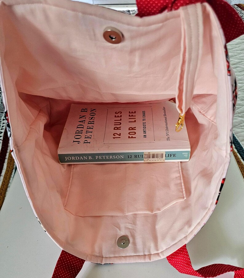 Puede incluir: Bolso tote abierto con interior rosa claro y un libro titulado "12 Reglas para la Vida" en su interior. El bolso tiene una correa de lunares rojos y un cierre magn&eacute;tico. El exterior presenta un dise&ntilde;o estampado.