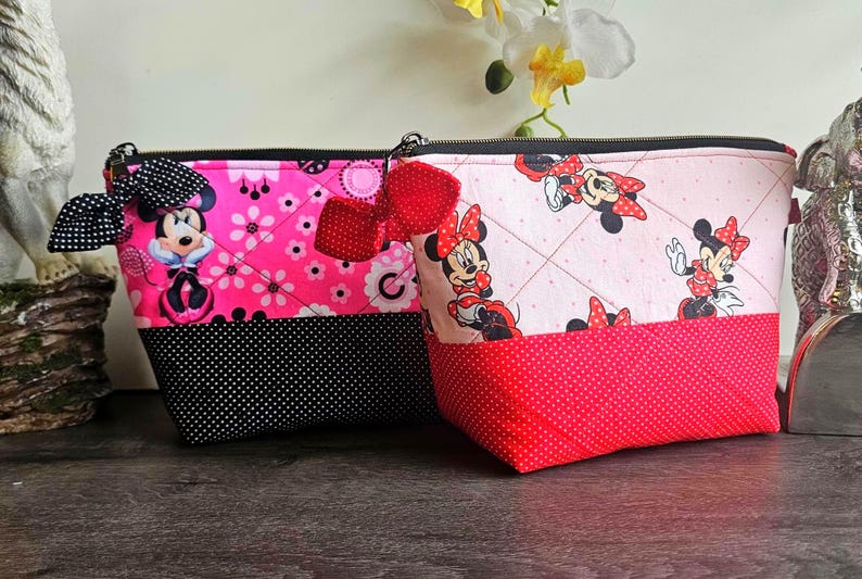 Peut inclure: Deux pochettes zipp&eacute;es matelass&eacute;es avec des motifs Minnie Mouse. La pochette de gauche est rose vif avec une base &agrave; pois noirs et un n&oelig;ud noir. Celle de droite est rose clair avec une base &agrave; pois rouges et un n&oelig;ud rouge. Fermetures noires.