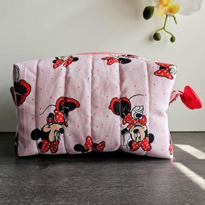 Puede incluir: Un neceser acolchado rosa con estampado de Minnie Mouse, lazos rojos y lunares. La bolsa tiene cierre de cremallera y una leng&uuml;eta roja en forma de lazo. La bolsa mide aproximadamente 20 cm de ancho y 15 cm de alto.