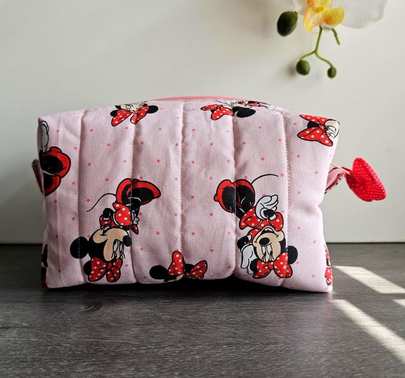 Puede incluir: Un neceser acolchado rosa con estampado de Minnie Mouse, lazos rojos y lunares. La bolsa tiene cierre de cremallera y una leng&uuml;eta roja en forma de lazo. La bolsa mide aproximadamente 20 cm de ancho y 15 cm de alto.