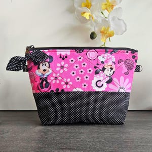 Peut inclure: Une pochette zipp&eacute;e matelass&eacute;e avec une section sup&eacute;rieure en tissu rose &agrave; motif Minnie Mouse et fleurs, et une section inf&eacute;rieure en tissu noir &agrave; pois blancs. Un n&oelig;ud en tissu &agrave; pois noirs et blancs est attach&eacute; &agrave; la fermeture &eacute;clair lat&eacute;rale.