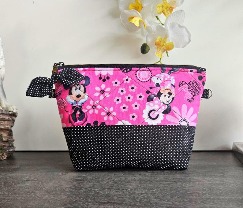 Peut inclure: Une pochette zipp&eacute;e matelass&eacute;e avec une section sup&eacute;rieure en tissu rose &agrave; motif Minnie Mouse et fleurs, et une section inf&eacute;rieure en tissu noir &agrave; pois blancs. Un n&oelig;ud en tissu &agrave; pois noirs et blancs est attach&eacute; &agrave; la fermeture &eacute;clair lat&eacute;rale.