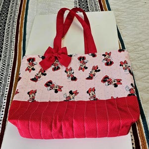 Puede incluir: Bolso tote acolchado con un estampado de Minnie Mouse sobre un fondo rosa claro. El bolso tiene asas rojas y un lazo rojo. La parte inferior del bolso es roja con un estampado de lunares.