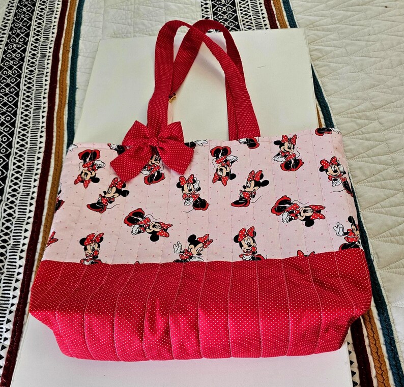 Puede incluir: Bolso tote acolchado con un estampado de Minnie Mouse sobre un fondo rosa claro. El bolso tiene asas rojas y un lazo rojo. La parte inferior del bolso es roja con un estampado de lunares.