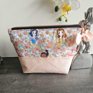 May include: A quilted zipper pouch featuring a white fabric top with a colorful floral pattern and cartoon princess illustrations, paired with a solid light pink fabric bottom. The pouch includes a black zipper and a light pink fabric bow accent.