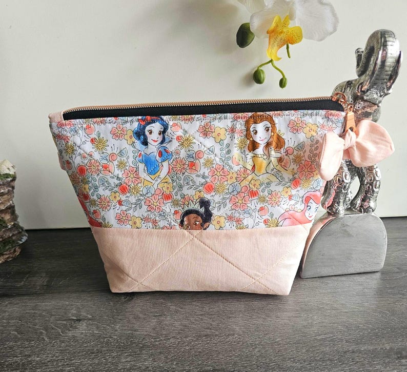May include: A quilted zipper pouch featuring a white fabric top with a colorful floral pattern and cartoon princess illustrations, paired with a solid light pink fabric bottom. The pouch includes a black zipper and a light pink fabric bow accent.