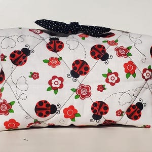 May include: White fabric bag with a ladybug and floral pattern. The bag features red ladybugs, red flowers, and green leaves on a white background. A black bow with white polka dots is on top.