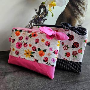 May include: Two quilted fabric pouches. One features a white background with ladybugs, flowers, and a pink bow zipper pull. The other has a white background with ladybugs, flowers, and a black bow zipper pull. Both have a pink bottom.
