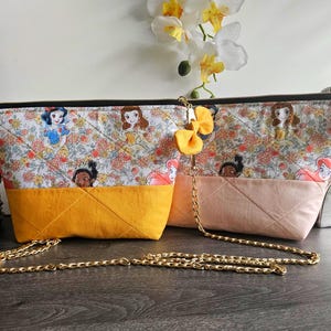 May include: Two quilted fabric pouches featuring a repeating pattern of cartoon princesses and floral motifs. One pouch has a mustard yellow base, the other a pale pink base. Both include a gold-tone metal chain strap and a small yellow fabric bow.