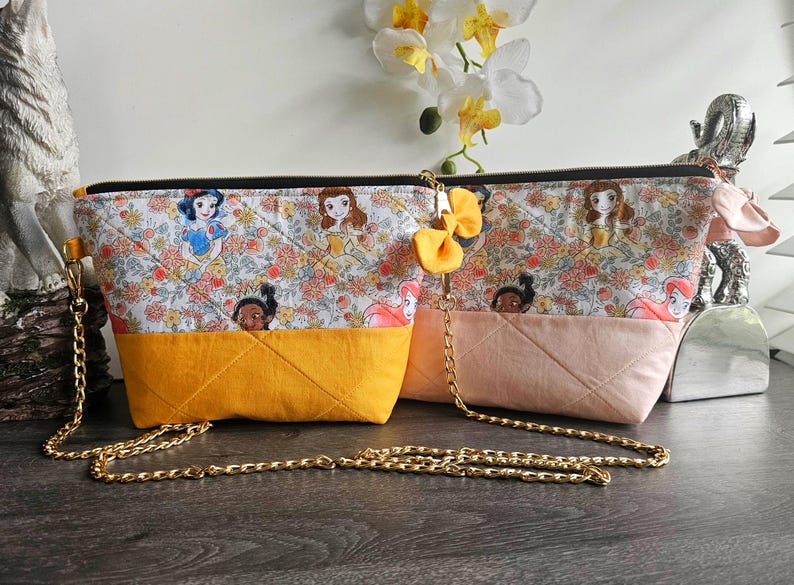 May include: Two quilted fabric pouches featuring a repeating pattern of cartoon princesses and floral motifs. One pouch has a mustard yellow base, the other a pale pink base. Both include a gold-tone metal chain strap and a small yellow fabric bow.