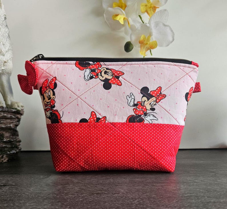 Pu&ograve; includere: Una pochette in tessuto trapuntato con un motivo ripetuto di Minnie Mouse su sfondo rosa chiaro a pois bianchi. La parte inferiore &egrave; di colore rosso tinta unita con pois bianchi. Include una chiusura a zip nera e un piccolo fiocco rosso laterale.