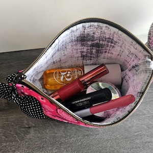 Peut inclure: Une pochette zipp&eacute;e rose avec un n&oelig;ud &agrave; pois noirs et blancs sur le c&ocirc;t&eacute;. L'int&eacute;rieur est doubl&eacute; d'un tissu blanc avec un motif noir vieilli. &Agrave; l'int&eacute;rieur se trouvent plusieurs articles cosm&eacute;tiques, dont un flacon dor&eacute; et divers tubes.
