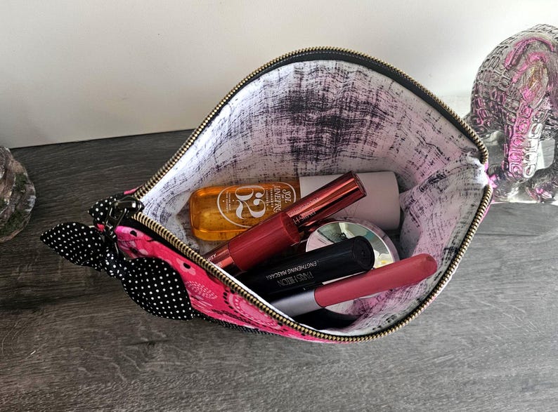 Peut inclure: Une pochette zipp&eacute;e rose avec un n&oelig;ud &agrave; pois noirs et blancs sur le c&ocirc;t&eacute;. L'int&eacute;rieur est doubl&eacute; d'un tissu blanc avec un motif noir vieilli. &Agrave; l'int&eacute;rieur se trouvent plusieurs articles cosm&eacute;tiques, dont un flacon dor&eacute; et divers tubes.