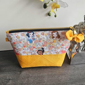 May include: A quilted zippered pouch featuring a white fabric top with a colorful print of various cartoon princesses and flowers, paired with a solid mustard yellow fabric bottom. The pouch includes a black zipper and a yellow fabric bow charm.
