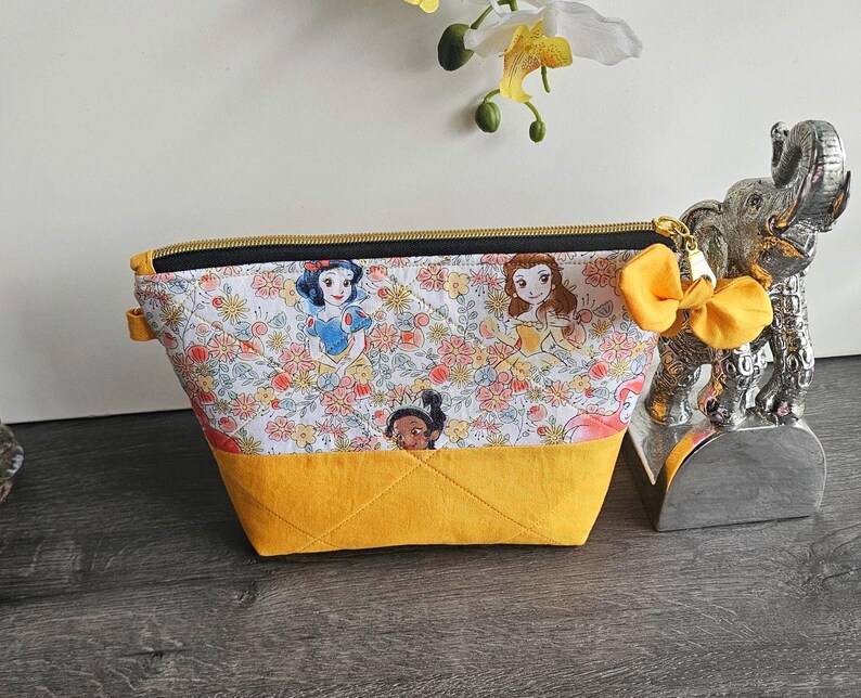May include: A quilted zippered pouch featuring a white fabric top with a colorful print of various cartoon princesses and flowers, paired with a solid mustard yellow fabric bottom. The pouch includes a black zipper and a yellow fabric bow charm.