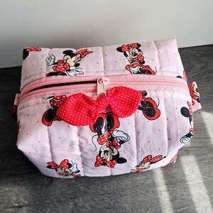 Puede incluir: Bolsa de cosm&eacute;ticos acolchada rosa con un estampado de Minnie Mouse, lazos rojos y lunares. Tiene una cremallera rosa y un lazo rojo decorativo. La bolsa rectangular es de tela.
