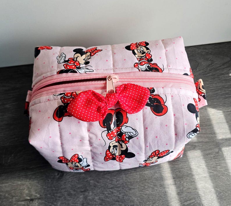 Puede incluir: Bolsa de cosm&eacute;ticos acolchada rosa con un estampado de Minnie Mouse, lazos rojos y lunares. Tiene una cremallera rosa y un lazo rojo decorativo. La bolsa rectangular es de tela.