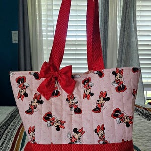 Puede incluir: Un bolso tote acolchado con una tela rosa claro estampada con Minnie Mouse. Tiene detalles de lunares rojos, un lazo grande y correas rojas. El bolso est&aacute; dise&ntilde;ado para transportar objetos.