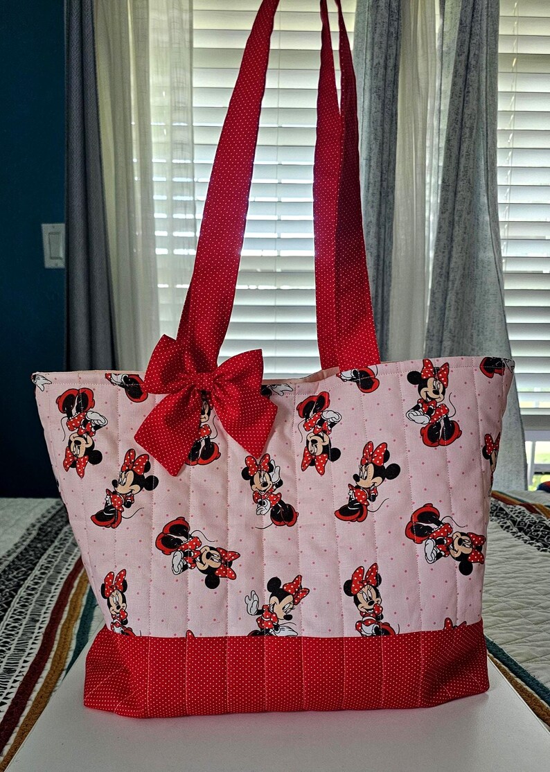 Puede incluir: Un bolso tote acolchado con una tela rosa claro estampada con Minnie Mouse. Tiene detalles de lunares rojos, un lazo grande y correas rojas. El bolso est&aacute; dise&ntilde;ado para transportar objetos.