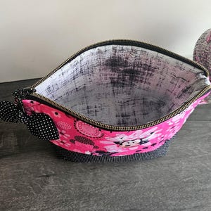 Peut inclure: Une pochette zipp&eacute;e avec un ext&eacute;rieur rose vif &agrave; motif Minnie Mouse, une base &agrave; pois noirs et blancs, et un n&oelig;ud &agrave; pois noirs et blancs attach&eacute; &agrave; la tirette. L'int&eacute;rieur est doubl&eacute; d'un tissu blanc pr&eacute;sentant une texture noire vieillie.