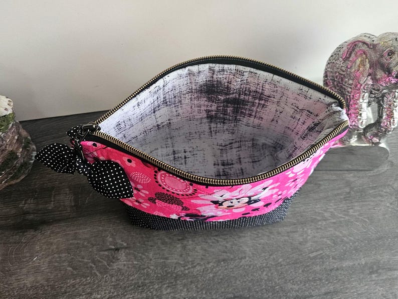 Peut inclure: Une pochette zipp&eacute;e avec un ext&eacute;rieur rose vif &agrave; motif Minnie Mouse, une base &agrave; pois noirs et blancs, et un n&oelig;ud &agrave; pois noirs et blancs attach&eacute; &agrave; la tirette. L'int&eacute;rieur est doubl&eacute; d'un tissu blanc pr&eacute;sentant une texture noire vieillie.