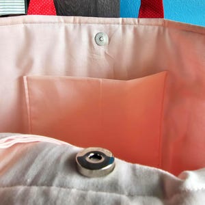 Puede incluir: Primer plano del interior de un bolso tote rosa. El bolso tiene un forro rosa claro con un peque&ntilde;o bolsillo. El borde superior del bolso tiene un cierre magn&eacute;tico a presi&oacute;n. Las asas son rojas con lunares blancos.