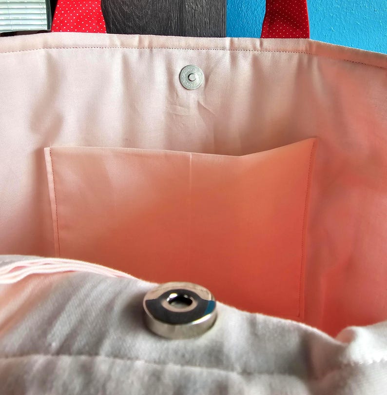 Puede incluir: Primer plano del interior de un bolso tote rosa. El bolso tiene un forro rosa claro con un peque&ntilde;o bolsillo. El borde superior del bolso tiene un cierre magn&eacute;tico a presi&oacute;n. Las asas son rojas con lunares blancos.