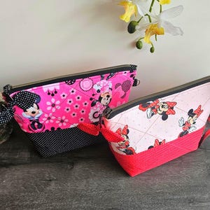 Peut inclure: Deux pochettes en tissu matelass&eacute; avec des motifs Minnie Mouse. La plus grande est rose vif avec une base &agrave; pois noirs et un n&oelig;ud noir. La plus petite est rose clair avec une base &agrave; pois rouges et un n&oelig;ud rouge. Toutes deux ont des fermetures &eacute;clair noires.