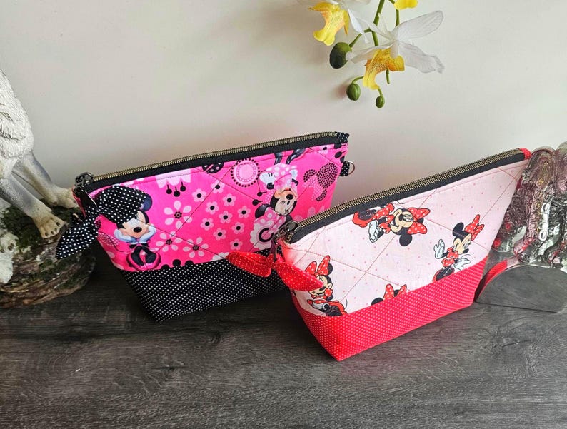 Peut inclure: Deux pochettes en tissu matelass&eacute; avec des motifs Minnie Mouse. La plus grande est rose vif avec une base &agrave; pois noirs et un n&oelig;ud noir. La plus petite est rose clair avec une base &agrave; pois rouges et un n&oelig;ud rouge. Toutes deux ont des fermetures &eacute;clair noires.