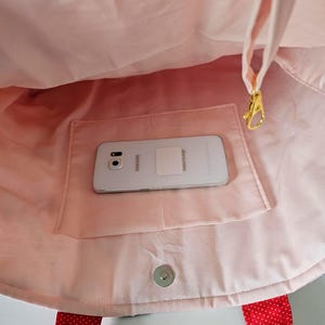 Puede incluir: Bolso tote rosa con asa de lunares rojos y un bolsillo. Un tel&eacute;fono Samsung Galaxy plateado descansa dentro del bolsillo. El bolso tiene un cierre magn&eacute;tico a presi&oacute;n y un cierre dorado.